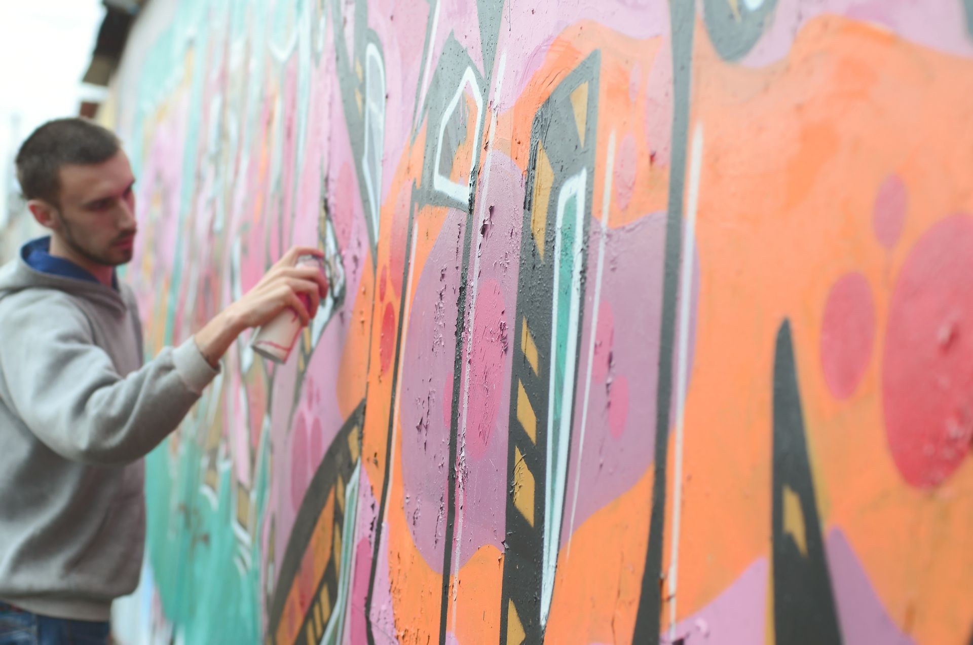 A young guy in a gray hoodie paints graffiti in pink and green c